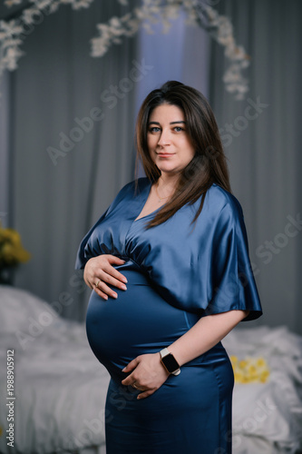 Portrait of pregnant woman in blue satin dress holding her belly, standing in soft lit bedroom, calm and confident expression, cozy home atmosphere, concept of motherhood, prenatal care and family