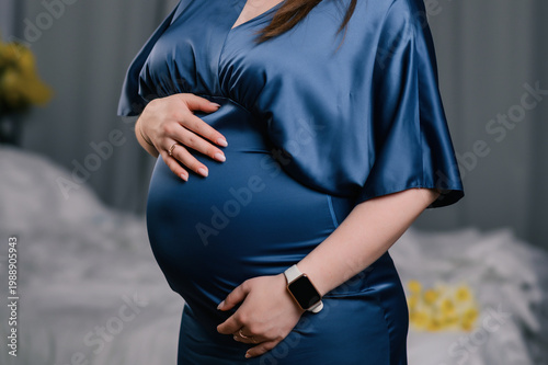 Close-up of pregnant woman in blue satin dress gently holding her belly, soft indoor lighting, cozy bedroom background, calm and caring mood, concept of motherhood, prenatal care and family lifestyle