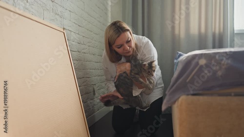 Woman greets curious cat near box gentle morning light bathes bedroom, exposed brick wall and soft curtain provide cozy backdrop, woman kneels to check box contents, lifts cat gently, pets and coax