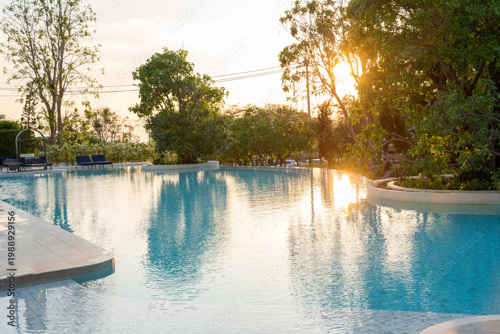 Obraz premium Luxury tropical resort swimming pool reflecting beautiful golden hour sunset light through green trees. Relaxing summer vacation, elegant hotel lifestyle, and peaceful nature travel getaway concept.
