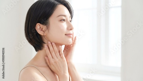 明るい窓辺のやさしい光の中でフェイスラインから首元に手を添え、美しい横顔となめらかな肌印象を見せる若い女性のビューティーイメージ、フェイスケア・ネックケア・美容広告に適した上品で清潔感のある人物素材