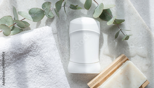 Fresh white deodorant stick surrounded by eucalyptus leaves towel and soap on light surface.