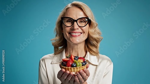 A woman in a white shirt and black glasses happily holding colorful vitamins in her hands in front of her face in a studio with a blue background.