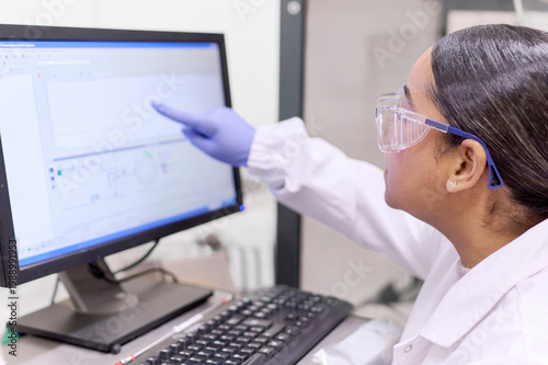 Female scientist with white coat and blue gloves observes and points to data on the laboratory computer where she works