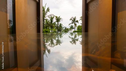 Photo of a symmetrical tropical resort glass table with a reflective surface that reflects palm trees and a cloudy sky, creating a calm, minimalist and luxurious holiday atmosphere.