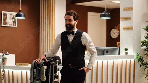 Vászonkép Cheerful smiling professional bellboy in formal attire with baggage trolley in front of check in reception
