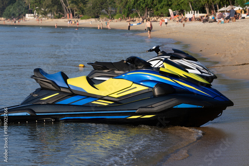 Fényképezés Two colorful jet skis rest on a tropical beach partly in water, with people swimming and relaxing under palm trees in the background
