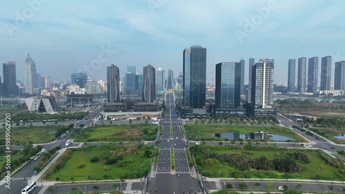 Wide view of developing district with symmetrical boulevard leading toward modern skyline and bridge, surrounded by green plots under cloudy daylight.
