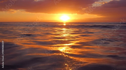 Sunset over calm ocean waves reflecting vibrant orange hues of setting sun on water surface with cloudy sky
