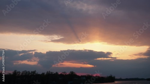 Clouds float over a lake as the sun sets in the background. Shadows form on the water. The colors change as daylight fades into night. Trees line the shore. Volume light atmosphere.