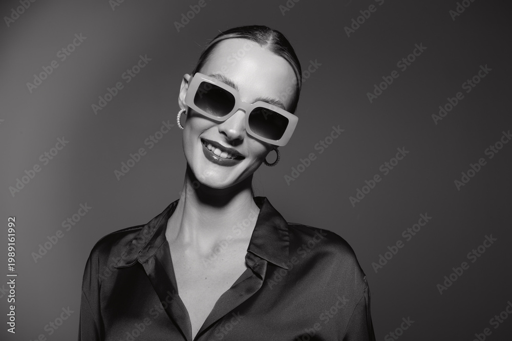 Obraz premium Smiling Caucasian woman with pale skin wearing oversized sunglasses and hoop earrings poses confidently in a dark satin blouse on grey background. Fashion and style concept.