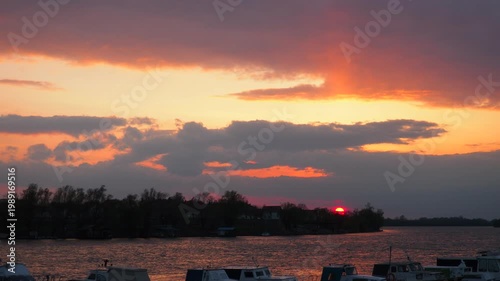 A colorful sunset is visible over a lake. Clouds reflect orange and purple hues. Boats are anchored in the harbor. The sun sets slowly behind the horizon.