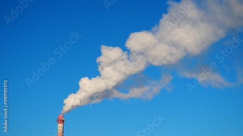 Factory chimneys release thick plumes of smoke into the air. The sky is clear and blue, with a few white clouds. This takes place in the afternoon, showcasing industrial activity.