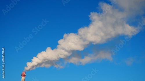 A factory chimney emits thick smoke into the sky. The clear blue background shows the contrast between the smoke and the environment. This happens in an industrial zone during the day.