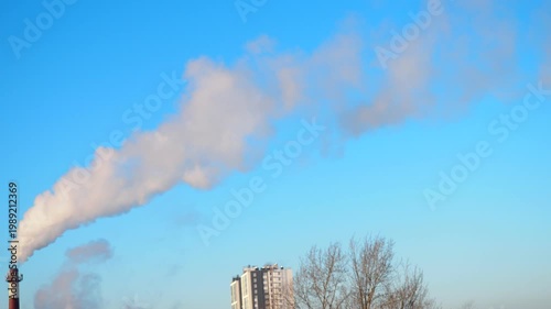 Smoke billows from a tall factory chimney as clear blue sky surrounds it. The activity takes place in an industrial area during late afternoon.