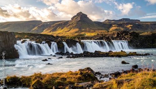 Waterfall cascades down rocky landscape with lush green grass and mountains in the background under a cloudy sky