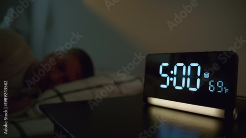 Man turning off alarm clock at 5,00 in bedroom. Early morning wake up moment with digital clock beside bed, 
