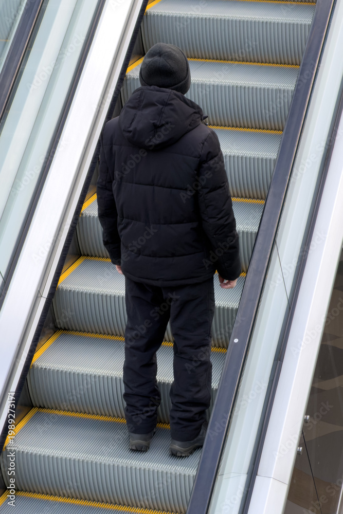 Fototapeta premium Back view of person on escalator, ascending indoors, wearing dark clothing
