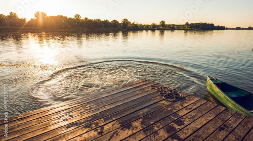 Konstfotografi Golden hour on a serene lake with a rustic wooden dock, a green boat, and a refreshing water splash