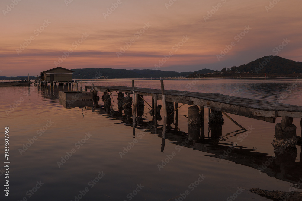 Obraz premium Wooden pier leading to a calm lake at sunset with warm orange sky, peaceful water reflections and distant hills, tranquil nature landscape perfect for travel, relaxation, serenity and scenic backgroun