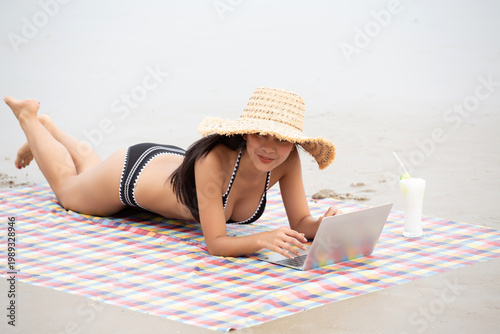 Obraz na plátně Southeast Asian woman relaxes on a sandy beach, enjoying a vibrant cocktail and a checkered blanket, wearing a black bikini and sunglasses, capturing a serene coastal moment