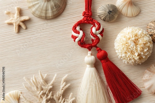 A flat lay composition showcases a decorative red and white braided tassel alongside a collection of seashells, starfish, and coral on a light wooden surface.