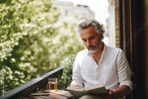 Focused middle-aged Hispanic man reads a book on a balcony on a sunny day