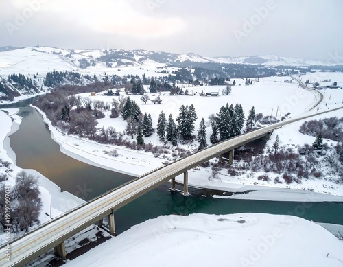Aerial View Of A Long Bridge Crossing A Frozen River In A Vast Snowy Winter Landscape With Distant Mountains And Pine Trees Under A Cloudy