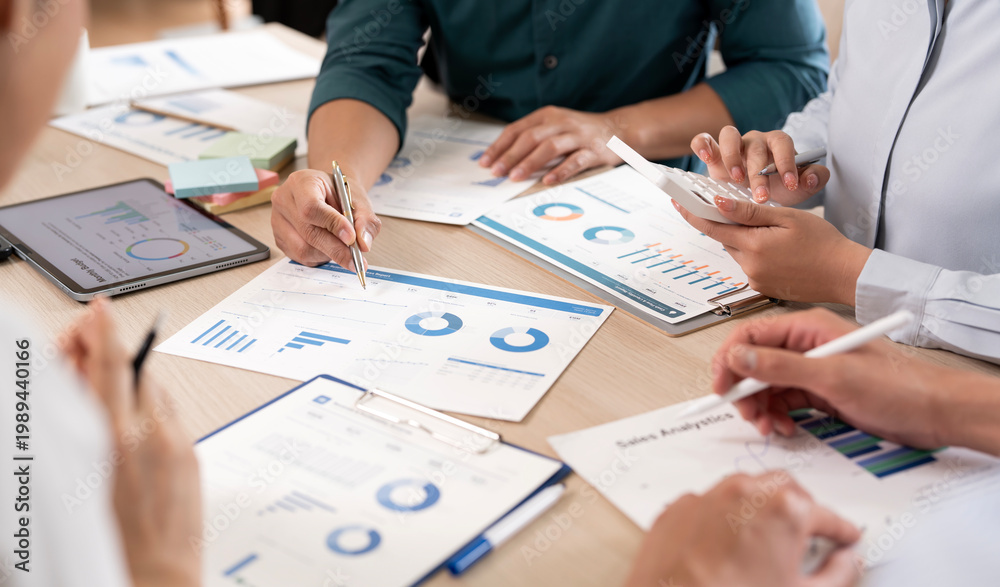 Obraz premium Professional business team collaborating on financial data and sales analytics using a calculator, tablet, and printed charts during a productive office meeting.