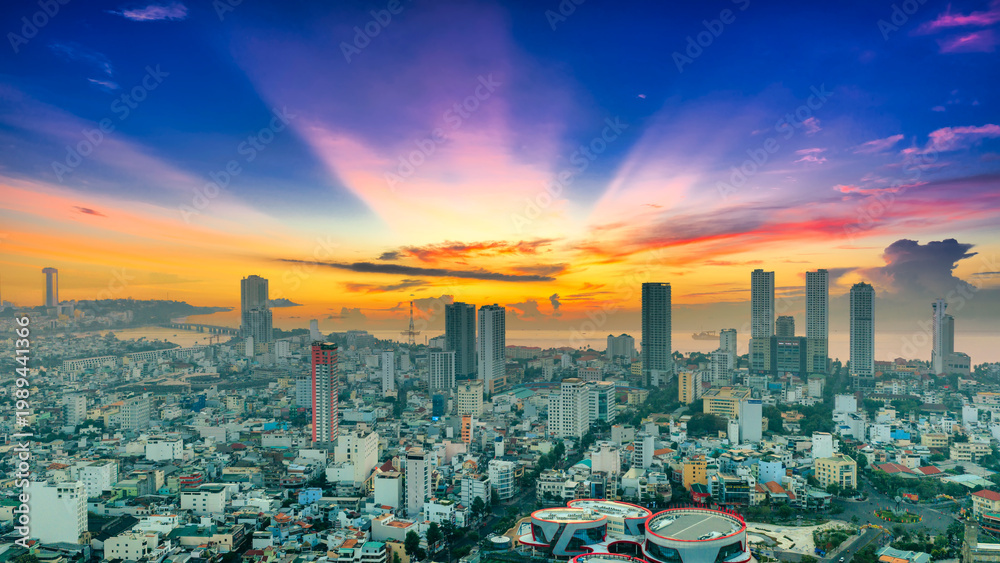 Obraz premium A serene sunrise view of Nha Trang city, Vietnam, where the soft orange sun rises above the coastal skyline. Layers of high-rise buildings and residential neighborhoods stretch toward