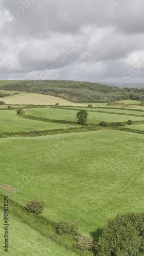 Rolling green fields and lush hedgerows stretch across a vibrant countryside landscape under a cloudy sky.