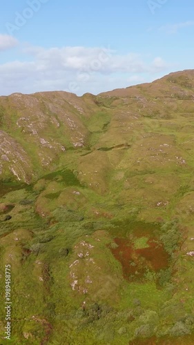 A wide aerial drone shot of green mountains and rolling hills dotted with rocky outcrops, all under a blue sky dotted with light clouds.