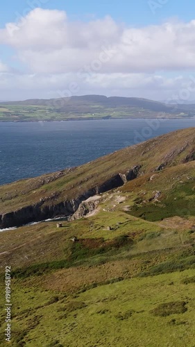 Sweeping aerial view of expansive green hills and dramatic rocky cliffs bordering the vast Atlantic Ocean in West Cork Ireland.