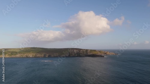 Capturing a vast coastal panorama with a verdant peninsula and rugged sea cliffs contrasting with the deep blue ocean. Bright sunlight illuminates the scene under a serene sky with white clouds.