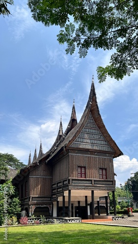 Traditional Indonesian house with curved, pointed rooflines rises against a bright blue sky,  features intricate decorative patterns on its wooden exterior and stands elevated on pillars.