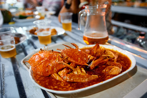 Sticker Steaming chili crab served in a rich red sauce on a communal table, surrounded by plastic cups of beer and casual diners enjoying a lively outdoor seafood meal