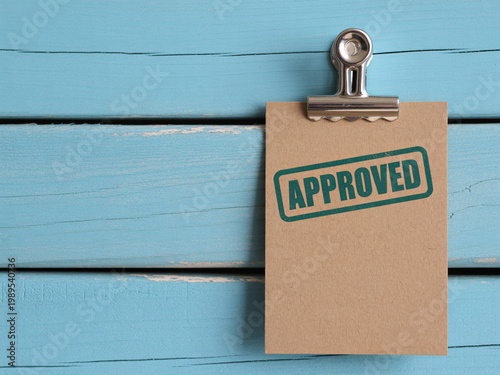 A clipboard with a piece of paper that has an approved stamp on it, hanging on a blue wooden wall.