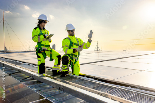 Two engineers in safety clothing with helmet check solar system at rooftop solar farm with city view. Engineers installing solar panels. Man and woman workers inspecting solar panels on roof at sunset