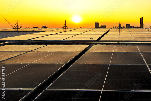 Photovoltaic solar panels on rooftop with a sunset over a solar farm. Solar panels installed on a building roof with city view generate clean, eco-friendly electricity. Producing renewable energy