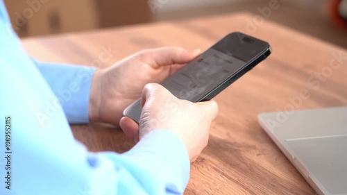 Close-up shows hands holding smartphone. Person wears light blue shirt. Laptop rests beside device. Person concentrates on screen content. Ideal for business or work scenarios