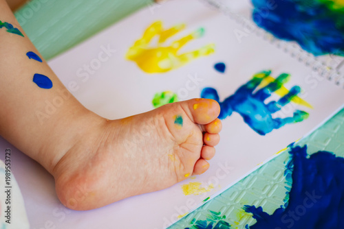 Baby paints on mat, tiny hands exploring blue green paint, colorful handprints and footprints, small paint pot tipped, playful messy texture, soft natural