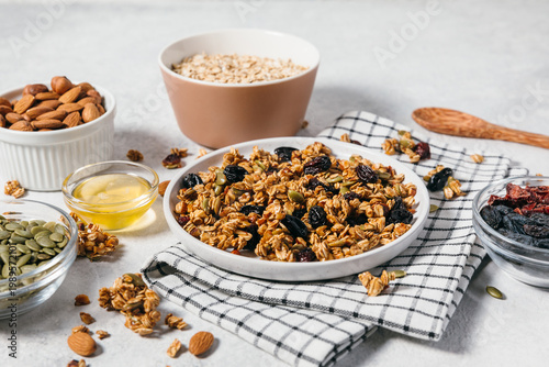 Crunchy organic granola clusters served on a plate and surrounded by raw ingredients like oats, nuts, and honey. Healthy breakfast, wholesome eating, homemade granola concept.