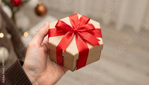 Hand holds gift box tied with red ribbon. Soft lighting highlights festive detail. Christmas tree ornaments visible in background. Warm, cozy holiday atmosphere. Focus on gift presentation
