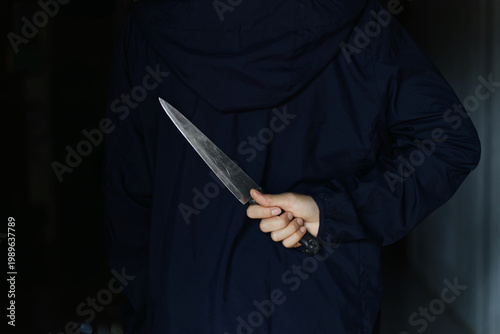 The Hidden Threat: Person in Dark Hoodie Concealing a Large Knife Behind Back