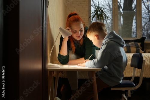 Mother helping her young son with homework at home under warm desk lamp light. Family support, education, parenting and focused study atmosphere. High quality photo