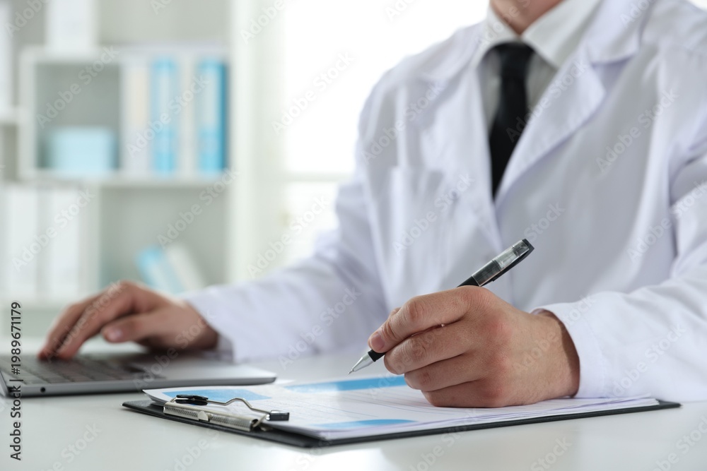 Fototapeta premium Doctor in medical coat working with clipboard and laptop at white table in clinic, closeup