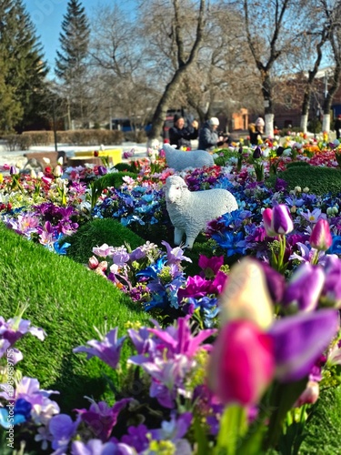 Decorative sheep statues on a vibrant multi-colored flower bed in a spring park.