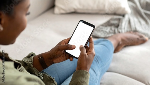 An individual, with dark skin, reclines comfortably on a sofa, casually holding a smartphone with a blank screen. The hands and bare feet are visible