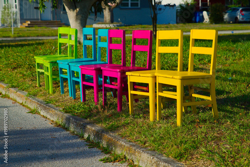 A row of vibrant painted wooden chairs in yellow, pink, blue and green sits on grass beside a curb, creating a playful, colorful outdoor seating display that evokes summer, community, and fun.
