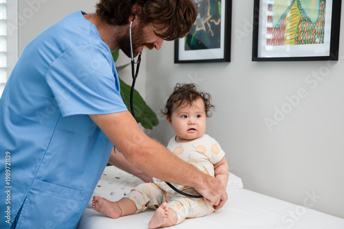 Photos Friendly male pediatrician in blue scrubs examining an infant with a stethoscope